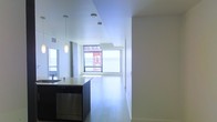 This single-floor space showcases a sleek, contemporary design characterized by an open-plan layout that seamlessly connects the kitchen and living areas. The kitchen is equipped with a dark-toned island unit, stainless steel dishwasher, and a light-colored countertop, complemented by a stylish pendant light that adds a focused decorative element overhead. The flooring throughout is a light-toned hardwood, contributing to the clean, minimalist feel of the interior. Large floor-to-ceiling windows at the far end of the living area flood the space with abundant natural light while framing views of the surrounding urban environment. The white walls and white ceiling further enhance the brightness and sense of openness throughout. Recessed and flush-mount ceiling lighting supplements the natural light, ensuring the space remains well-lit at all times. A corridor extends toward a private area of the unit, suggesting additional rooms beyond the main living space. Overall, the space conveys a polished, modern ambiance with a neutral palette and well-considered architectural details.