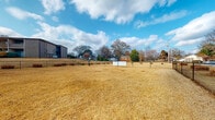 This community dog park offers a well-rounded set of amenities designed to serve both pets and their owners. The outdoor area is a generously sized, fully fenced open field where dogs can roam freely. The ground is largely open and level, bordered by mature trees that provide a natural backdrop, with neighboring structures visible along the perimeter. A gated entry section within the fencing provides a secure transition point for entering and exiting with pets.

On-site, a dedicated laundry facility serves the community with an extensive lineup of both top-loading and front-loading washers and dryers. The room features light wood-look flooring, ceiling fans for ventilation, and windows that allow natural light to filter in. A folding table provides a practical workspace, and posted laundry guidelines are clearly visible.

A small ancillary building adjacent to the park contains a compact, open interior with wood-look flooring, a work surface, and large windows offering direct views of the outdoor grounds. This space appears to function as a rest or management area, providing a comfortable vantage point overlooking the park. Together, these features create a functional and community-oriented environment.