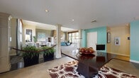 This single-floor community clubhouse offers a thoughtfully designed layout that seamlessly blends comfort and functionality across multiple distinct areas. The open-plan main lounge serves as the centerpiece, featuring a striking stacked-stone fireplace flanked by large artwork, a teal sofa, and plush seating arrangements set atop richly patterned carpet in deep burgundy and teal tones. White decorative columns define zones throughout the space without interrupting the open flow. A casual dining and gathering area showcases a dark wood table, decorative centerpiece, and bold teal accent walls that add a vibrant, contemporary touch. Lush potted plants are placed throughout, enhancing the welcoming ambiance. A dedicated private screening room is outfitted with rows of navy blue theater-style seating, film reel-patterned carpeting, and a classic red popcorn machine — ideal for movie screenings and events. An entrance foyer with large windows and French doors invites abundant natural light, offering views of the surrounding greenery. The kitchen features white cabinetry, dark countertops, and tile flooring, providing a practical space for catering and food preparation. Recessed lighting throughout maintains a warm, inviting atmosphere across all areas of this versatile community facility.