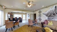 This single-story space serves as a comfortable communal lounge designed to accommodate both casual socializing and relaxed seating. The open layout is anchored by a central seating arrangement featuring a round table paired with plush upholstered chairs, ideal for small group gatherings. A textured area rug in warm golden tones grounds this central zone, adding warmth and visual interest to the space.

To one side, a raised bar-height seating area with dark wood stools and tall chairs offers an alternative setting for conversation or casual dining. The opposite side of the room features a leather seating arrangement, contributing a more formal lounge feel.

An arched passageway connects the main room to an adjoining area, which benefits from natural light streaming through large windows. Recessed lighting and a ceiling fan with an integrated light fixture provide layered illumination throughout the space. Crown molding along the upper walls adds a refined architectural detail.

Decorative wall art, including geometric metal accents and a stylized animal motif, lends a distinctive, curated character to the interior. The overall ambiance is inviting and versatile, balancing comfort with thoughtful design.