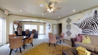 This single-story space serves as a comfortable communal lounge designed to accommodate both casual socializing and relaxed seating. The open layout is anchored by a central seating arrangement featuring a round table paired with plush upholstered chairs, ideal for small group gatherings. A textured area rug in warm golden tones grounds this central zone, adding warmth and visual interest to the space.

To one side, a raised bar-height seating area with dark wood stools and tall chairs offers an alternative setting for conversation or casual dining. The opposite side of the room features a leather seating arrangement, contributing a more formal lounge feel.

An arched passageway connects the main room to an adjoining area, which benefits from natural light streaming through large windows. Recessed lighting and a ceiling fan with an integrated light fixture provide layered illumination throughout the space. Crown molding along the upper walls adds a refined architectural detail.

Decorative wall art, including geometric metal accents and a stylized animal motif, lends a distinctive, curated character to the interior. The overall ambiance is inviting and versatile, balancing comfort with thoughtful design.