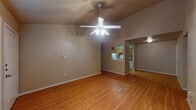 This single-floor unit is characterized by consistent warm-toned hardwood-style flooring that runs throughout the living spaces, bedrooms, kitchen, and bathroom, creating a cohesive and easy-to-maintain interior. The living room stands out with its vaulted ceiling height, lending the space an open and airy feel, and is equipped with a ceiling fan fitted with an integrated light fixture. A pass-through opening connects the living area to the galley-style kitchen, which features warm oak cabinetry on both sides, dark laminate countertops, a double-basin stainless steel sink, a white electric coil-burner range, a white refrigerator, a dishwasher, and overhead ventilation. The layout includes multiple bedrooms, each finished with the same hardwood-style flooring, neutral beige walls, white trim, and ceiling fans with light fixtures. Select bedrooms feature windows fitted with horizontal blinds, allowing for natural light control. The bathroom includes a full-size soaking tub with a tiled shower surround, chrome fixtures, a toilet, and a vanity with storage. Throughout, the neutral color palette and clean finishes provide a functional, move-in-ready environment.