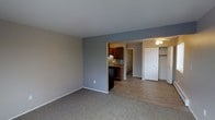 This single-floor space showcases an open-concept layout that seamlessly connects the main living area to the kitchen and bedroom. The living area is finished with neutral-toned carpet, while the kitchen and adjoining spaces transition to smooth laminate wood-look flooring. Walls are painted in a warm greige tone with a contrasting cool gray accent wall that defines the boundary between the living and kitchen areas, adding visual depth to the space.

The kitchen features rich, dark wood cabinetry paired with a decorative mosaic tile backsplash, offering a stylish focal point. A black appliance suite complements the cabinetry, creating a cohesive and practical cooking space.

Visible beyond the kitchen, the bedroom is equipped with a ceiling fan with integrated lighting, providing both comfort and illumination. A set of bi-fold closet doors reveals a spacious reach-in closet with a hanging rod, offering ample storage. White trim and baseboards throughout the unit provide a clean, finished look that ties each area together into a cohesive and functional living environment.