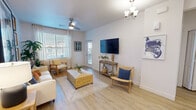 This well-designed single-floor residence offers a cohesive blend of modern aesthetics and comfortable living across its thoughtfully arranged spaces. The living room serves as the heart of the home, featuring light hardwood-style flooring, crisp white walls, and an abundance of natural light streaming through large windows dressed with sheer curtains and wood-slat blinds. A ceiling fan and elegant chandelier provide dual lighting options, while a mounted flat-screen television anchors the main wall above a rustic wood console table. The kitchen is sleek and functional, showcasing white shaker-style cabinetry, quartz countertops, stainless steel appliances including a gas range, and a peninsula breakfast bar illuminated by pendant lighting—ideal for casual dining or entertaining. Both bedrooms are serene and well-proportioned, each benefiting from natural light through shuttered windows and ceiling fans for year-round comfort. The bathroom features a wide vanity with a white quartz countertop, an undermount sink, a large wall mirror, and a tiled shower enclosure. Throughout the residence, light wood-tone flooring ties each space together, creating a warm, airy atmosphere that is both modern and inviting.
