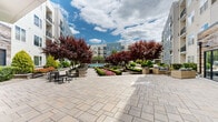 Located at 547 Old Bridge Tpk in East Brunswick, NJ, this beautifully designed outdoor courtyard serves as the central amenity space of a modern multi-story residential community. The grounds are thoughtfully landscaped, featuring striking deep-red ornamental trees, neatly trimmed boxwood hedges, vibrant seasonal flower beds in pink and white, and manicured green lawns that contribute to a lush, park-like atmosphere.

A spacious stone-paved promenade anchors the courtyard, leading residents from one end of the property to the other. At the heart of the space lies a resort-style swimming pool, enclosed by an iron fence and surrounded by stone accent walls with lush plantings. Adjacent to the pool, multiple seating zones offer varied social settings, including cushioned lounge chairs and wicker conversation sets ideal for relaxation or gathering.

Additional outdoor amenities include dining table sets, benches along garden pathways, and a dedicated grilling area with covered equipment. The surrounding multi-story buildings feature private balconies that overlook the courtyard, creating a cohesive sense of community. Ground-level units benefit from direct access to the landscaped lawn areas. The entire space is bathed in natural light, with open skies enhancing the inviting, resort-inspired ambiance throughout.