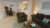 This single-floor residence offers a thoughtfully designed open-concept layout that seamlessly connects the living, dining, and kitchen areas. The living room is a welcoming focal point, furnished with plush, dark upholstered seating arranged around a patterned area rug and a wood coffee table, creating a cozy yet functional gathering space. Recessed lighting and neutral, warm-toned walls contribute to the bright and inviting atmosphere throughout.

The kitchen features crisp white cabinetry and connects to the main living area via a convenient pass-through counter, allowing for easy interaction between spaces. This layout is well-suited for both everyday living and entertaining. Adjacent to the living room, the dining area accommodates a dark wood table and chairs, providing a defined space for meals while remaining open to the rest of the home.

Consistent light-colored carpeting flows throughout the main living areas, adding warmth and continuity to the design. Crown molding along the ceilings lends an elegant architectural detail, elevating the overall aesthetic of the space. The single-floor configuration ensures effortless movement between rooms, making the layout both practical and comfortable for day-to-day living.