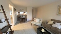 This single-floor space is designed with an open-concept layout that seamlessly connects the living area to the kitchen. The living room features neutral wall-to-wall carpeting and is furnished with a comfortable seating arrangement, centered around a dark coffee table and an accent loveseat with a floral pattern, adding a touch of character to the space. The walls are painted in a soft, warm neutral tone that enhances the bright, airy feel throughout.

The kitchen is positioned at the back of the open layout and is partially separated by a raised breakfast bar, which serves as both a functional dining surface and a visual divider between the two spaces. The bar accommodates seating for multiple guests with the inclusion of wooden bar stools. Dark cabinetry lines the kitchen walls, providing ample storage, and a window above the counter allows natural light to filter into the cooking area. Decorative metal lettering on the wall above the kitchen adds a personalized, welcoming touch.

A hallway visible from the living area leads to additional spaces within the unit. The overall ambiance is casual and functional, with a layout well-suited for everyday living and entertaining.