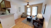 This single-floor residence offers a warm and inviting atmosphere, thoughtfully laid out to maximize comfort and functionality. The living area serves as the heart of the home, featuring smooth hardwood-style flooring that runs throughout and neutral-toned walls that create a bright, airy feel. Two windows dressed with horizontal blinds and teal curtain panels allow generous natural light to filter into the room, enhancing the open ambiance. A ceiling fan with a light fixture provides both airflow and overhead illumination, contributing to the space's practical charm. The living room is well-proportioned, comfortably accommodating a dark leather sofa and a reclining armchair arranged around a tufted ottoman that doubles as a coffee table, all anchored by a patterned area rug. A floor lamp adds warm ambient lighting to one corner of the room. Adjacent to the living area, a partial view of the kitchen reveals a countertop with a light granite finish, hinting at additional functional space nearby. The front entry door is conveniently located within the living area, making for a seamless transition from the exterior into the home's welcoming interior. Overall, the space presents a cozy, move-in-ready environment with a clean, casual aesthetic.
