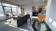 This contemporary urban residence is thoughtfully designed across a single floor, blending industrial and modern aesthetics to create a stylish and functional living environment. The open-plan layout seamlessly connects the living, dining, and kitchen areas, fostering a sense of spaciousness and flow throughout.

The living area is anchored by a large, dark sectional sofa and features dramatic floor-to-ceiling windows that flood the space with natural light and frame expansive city views. The exposed concrete ceiling adds an industrial character that complements the modern furnishings. Sleek accent chairs and carefully placed décor contribute to the room's contemporary feel.

Adjacent to the living area, the dining space features a glass-top table paired with modern white chairs, creating a clean and minimalist aesthetic. A round side table with a decorative glass bowl centerpiece adds a refined touch.

The kitchen is defined by a warm amber-toned island counter that introduces a bold pop of color against the otherwise neutral palette. It opens directly to the main living area, making it ideal for entertaining. Beyond the kitchen, a hallway leads to additional private spaces within the residence.

Throughout, the interplay of natural light, urban views, and a cohesive modern design palette makes this space a compelling and well-appointed urban retreat.