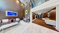 Located at 717 Preakness Avenue in Totowa, New Jersey, this sophisticated building lobby makes a striking first impression with its dramatic double-height ceilings and carefully curated design elements. The main entry area showcases light-toned, large-format porcelain tile flooring that contrasts beautifully against the rich, warm brown walls, creating a refined and welcoming atmosphere.

A towering stacked-stone accent wall serves as the lobby's focal centerpiece, adorned with a decorative floral wreath that adds an elegant, organic touch. Luxurious tufted gray velvet seating arrangements are thoughtfully positioned throughout the space, complemented by a crisp white coffee table and curated decorative accessories. Framed artwork enhances the walls, contributing to the upscale aesthetic.

The lobby flows seamlessly into a carpeted corridor area, where a richly patterned brown carpet with floral motifs guides visitors toward the building's elevator — finished in sleek stainless steel. This secondary corridor is illuminated by a stunning fringe chandelier and warm recessed lighting, maintaining the design continuity established in the main entry.

Crown molding, decorative wall paneling, and layered lighting — including an eye-catching sculptural ceiling fixture — elevate the space further. A mezzanine-level glass railing is visible above, adding architectural depth. Overall, the lobby presents a polished, hotel-inspired environment that reflects meticulous attention to detail and design.