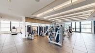 Located at 8500 N Sherman Circle in Miramar, Florida, this well-appointed fitness center offers a generously sized, single-floor exercise space designed to support a full range of workout activities. The layout is thoughtfully organized into distinct zones, including a cardio area lined with treadmills, ellipticals, and stationary bikes, and a strength training section equipped with a variety of cable and resistance machines. A glass-enclosed studio room provides a dedicated space for free weights, benches, and focused training sessions, with wall-to-wall mirrors and mounted flat-screen displays enhancing functionality. The flooring transitions between durable rubber sport tiles in the main workout areas and light hardwood in the studio, offering both practicality and visual appeal. A dropped ceiling with recessed linear lighting runs throughout, creating a clean, contemporary atmosphere. Textured white stone accent walls add an architectural element, while large windows along the perimeter invite natural light and views of the surrounding greenery. The space also features a corridor with glass balustrade railings and additional wall-mounted TVs, contributing to a polished, modern environment well-suited for fitness and wellness activities.