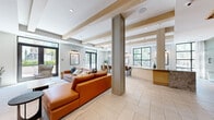 This thoughtfully designed single-floor space in Walnut Creek, California, offers a generously proportioned open-concept layout that seamlessly integrates multiple functional areas. The interior is defined by exposed wood beam detailing across the ceiling, adding warmth and architectural character to the bright, airy environment.

Large black-framed windows and glass doors line the perimeter, flooding the space with natural light and providing views of the surrounding outdoor greenery. The flooring throughout consists of large-format light stone tiles that contribute to a clean, cohesive aesthetic.

The lounge area features a substantial leather sectional sofa in a rich cognac tone, complemented by a striking stone-clad feature wall with a built-in wood-framed media niche. Adjacent to the lounge, a dramatic oversized marble-topped island serves as a central gathering point, surrounded by upholstered counter-height seating and illuminated by a series of elegant pendant lights in varying styles.

A kitchenette area with cabinetry and shelving supports the space's hospitality function, while a wood-slatted accent wall adds texture near a more intimate seating nook. The overall ambiance is modern yet welcoming, balancing refined materials with a comfortable, community-oriented atmosphere.