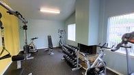 This fitness space is designed to support a well-rounded workout routine within a compact, functional layout. The room features light sage-green walls and a textured ceiling with flush fluorescent lighting, creating a clean and practical environment. Rubber flooring throughout provides durability and safety underfoot during exercise. The equipment selection is comprehensive, including a stationary cycling bike, an elliptical trainer, and an additional cardio machine, offering a variety of cardiovascular workout options. A adjustable weight bench sits centrally, complemented by a well-organized rack of dumbbells ranging across multiple weight increments, making strength training accessible for various fitness levels. A notable architectural feature is the brick fireplace situated in the corner of the room, adding a distinctive character element that contrasts with the otherwise utilitarian design. A window on one wall allows for a degree of natural light to enter the space. The single-floor layout ensures easy accessibility, and the thoughtful arrangement of equipment maximizes the available floor area without feeling overcrowded, making this a practical and inviting fitness facility.