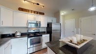 Located in Dublin, Ohio, this single-floor residence offers a thoughtfully designed layout that balances functionality with comfort. The kitchen is a standout feature, showcasing crisp white shaker-style cabinetry, dark granite countertops, hardwood-style flooring, and a full suite of stainless steel appliances including a range with over-the-range microwave, refrigerator, and dishwasher. A rustic wooden accent beam above the cabinetry adds a charming contrast to the clean, modern finishes throughout.

The living area is generously proportioned, with plush carpeting, neutral-toned walls, and a large sliding glass door that invites an abundance of natural light and provides a view of the outdoors. Track lighting overhead adds a contemporary touch to the open and airy atmosphere.

The bedroom is a peaceful retreat, featuring soft carpeting, a corner window dressed with elegant draping curtains, and direct access to the en-suite bathroom. The bathroom is particularly well-appointed, offering a double-sink vanity with granite countertops and white cabinetry, a walk-in glass shower with tile surround, and ample storage. The cohesive use of neutral tones and quality finishes throughout the home creates a warm, inviting ambiance ideal for comfortable everyday living.