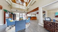 This single-story clubhouse facility offers a thoughtfully designed community space that blends modern aesthetics with warm, inviting materials throughout. The grand reception lobby immediately impresses with soaring ceilings, a distinctive coffered wood-paneled ceiling detail, and elegant globe pendant lighting. Sleek hardwood-style flooring runs throughout, while a seating area furnished with blue accent chairs and a colorful area rug creates a welcoming atmosphere for visitors. A warm wood-paneled reception desk anchors the space, complemented by curated artwork and display shelving with integrated media screens. Large glass doors and clerestory windows flood the lobby with natural light and provide views to the outdoors. Adjacent to the lobby, a well-appointed games room offers ample entertainment options, including a pool table, shuffleboard table, arcade machine, and a lounge area set against a bold blue feature wall with vibrant artwork. Exposed wood beam accents add architectural character throughout this space. The fitness center is generously sized and well-lit, featuring large windows that bring in natural light and outdoor views. It is outfitted with treadmills, ellipticals, cycling bikes, cable machines, free weights, and a ceiling fan, all set atop durable hardwood-style flooring with full-length mirrors enhancing the sense of space.