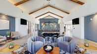 Located at 5700 Crossings Blvd in Nashville, TN, this single-story community clubhouse offers a thoughtfully designed, expansive gathering space spanning a generous floor area. The interior makes an immediate impression with soaring vaulted ceilings accented by exposed wood beams, polished concrete flooring throughout, and a cohesive blue-and-neutral color palette that lends a sophisticated yet welcoming atmosphere.

The reception area greets visitors with warm wood cabinetry, recessed and pendant lighting, and curated greenery. Adjacent lounge zones feature plush, channel-tufted seating in muted blues and grays, layered over bold patterned rugs. A dramatic floor-to-ceiling fireplace wall clad in large-format slate-toned panels serves as a striking focal point, flanked by rounded lounge chairs and sculptural pendant fixtures.

The central great room showcases the full height of the vaulted ceiling with exposed beams, leading to a stylish bar area highlighted by a blue tile backsplash, pendant lighting, and bar seating alongside a communal dining table. Floor-to-ceiling steel-framed windows flood the space with natural light and provide views to an outdoor terrace. Decorative wall art and contemporary sconce lighting complete the polished, design-forward aesthetic throughout.