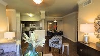 This single-floor residence showcases an open-concept layout that seamlessly connects the kitchen, dining, and living areas into one cohesive and functional space. Dark hardwood floors run throughout, providing a rich, warm contrast to the soft gray-toned walls accented by crisp white crown molding. A ceiling fan with a light fixture anchors the living area, contributing to the overall brightness of the space.

The kitchen features dark cabinetry paired with stainless steel appliances, including a refrigerator and microwave, and is separated from the main living area by a breakfast bar with light-colored countertops. Decorative columns with white capitals frame the transition between the kitchen and the adjoining dining area, adding an architectural element that enhances the sense of structure without disrupting the open feel.

The dining area is defined by a pendant light overhead and accommodates a modest dining set, creating a dedicated space for meals. The living area offers room for seating, highlighted by a patterned accent chair with geometric detailing. Together, the rooms create a cohesive, well-appointed interior with a clean, contemporary aesthetic.