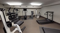 This fitness center offers a practical and functional workout environment outfitted with a diverse range of exercise equipment to accommodate various training needs. The space is laid out across a single floor, providing ample room to move between stations without feeling cramped.

Cardio equipment is well represented, with multiple treadmills, a recumbent stationary bike, an upright bike, and an elliptical trainer positioned throughout the room. A wall-mounted television is conveniently located near the treadmills, allowing users to stay entertained during cardio sessions.

Strength training is equally well served, with several cable and weight-stack machines designed for targeted muscle group exercises. These include units suited for leg work, upper body conditioning, and multi-function strength movements. Rubber floor mats are placed beneath select equipment to protect the carpeted flooring and provide stability during use.

The room features neutral-toned walls, textured ceilings, and recessed fluorescent lighting that ensures bright, even illumination throughout. Small windows allow for a degree of natural light to supplement the overhead fixtures. Ventilation is provided through ceiling-mounted HVAC vents. The overall atmosphere is clean, straightforward, and purpose-driven, making it a reliable facility for regular fitness use.