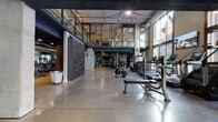 This two-floor fitness facility showcases a striking industrial design, characterized by exposed concrete ceilings, visible ductwork, steel-framed partitions, and polished concrete floors throughout the main level. The ground floor is a generously proportioned open workout space bathed in natural light from a tall bank of windows along one wall, providing views of the surrounding streetscape. A standout feature of this level is a built-in rock climbing wall, offering an unconventional training option alongside a full array of cardio machines including treadmills and ellipticals, resistance and cable equipment, free weights, and benches. Large mirrors line sections of the walls, amplifying the sense of space. A dedicated free weight area with darker overhead steel decking creates a distinct zone within the floor.

The upper level, accessible via an open staircase with industrial metal railings, transitions to warm hardwood flooring and houses additional strength and functional training equipment, including multi-station cable rigs and battle ropes. A mirrored studio space on this level features hardwood floors, track lighting, and open sightlines to the floor below through wire-mesh railings, making it well-suited for group fitness or personal training sessions. Throughout both floors, the raw architectural details lend the facility a dynamic, urban character.
