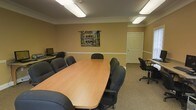 This single-floor space is designed to serve as a functional and comfortable meeting environment. The conference room is anchored by a large, oval-shaped wooden table surrounded by multiple ergonomic rolling chairs, providing ample seating for group meetings, presentations, or collaborative work sessions. The room is finished with warm, neutral-toned walls accented by classic crown molding, adding a touch of architectural detail. Overhead, fluorescent lighting fixtures ensure consistent, even illumination throughout the space. A decorative tapestry depicting a street scene hangs on the wall, lending a touch of character to the room. Along one wall, a compact workstation equipped with a desktop computer offers additional utility for tasks requiring individual focus. A secondary desk and chair area near the far corner provides further flexible workspace. The space is completed by a paneled door offering access to an adjacent area. Carpeted flooring in a neutral tone ties the room together and contributes to a quiet, professional atmosphere well-suited for focused discussions and productive meetings.