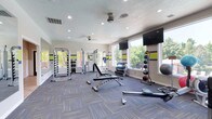 This fitness center offers a thoughtfully designed workout environment spread across a single floor. The space is flooded with natural light through large windows that line the walls, providing views of lush greenery outside and creating an open, motivating atmosphere. The interior is finished with patterned blue and grey carpet tiles and accented with bold yellow and blue horizontal stripes along the walls, lending the space a dynamic and energetic aesthetic. Recessed lighting and dual ceiling fans ensure the area remains well-lit and comfortable during exercise sessions. The facility is comprehensively equipped to support a wide range of training styles. Strength training is well-served by a multi-function cable machine, a weight rack stocked with a full set of dumbbells, and an adjustable weight bench. For functional and cardio training, the space includes medicine ball racks, a rowing machine, and additional floor space for stretching or bodyweight exercises. Multiple wall-mounted flat-screen televisions are positioned throughout the room, enhancing the workout experience. The layout is spacious and uncluttered, allowing for comfortable movement between equipment stations. Overall, this fitness center combines practical functionality with a clean, modern design to deliver an effective and enjoyable workout environment.
