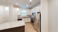 Located at 1000 Infinity Dr in Maryville, Tennessee, this single-floor residence offers a clean, modern aesthetic with consistent wood-look flooring and crisp white walls throughout. The open-concept kitchen is a standout feature, equipped with stainless steel appliances including a refrigerator, range, and microwave, complemented by white shaker-style cabinetry, granite countertops, and a dark-based center island with pendant lighting overhead. A window above the sink brings in natural light, enhancing the bright and functional layout. The kitchen flows seamlessly into the living area, which provides an open and airy feel. Each bedroom is generously sized, featuring ceiling fans with light fixtures and windows with blinds that allow for natural light and outdoor views. The primary bathroom includes a dual-sink vanity with granite countertops, white cabinetry, and a large mirror with overhead lighting. A walk-in closet is accessible directly from the bathroom, offering ample storage. Additional bathrooms follow the same refined design language. Clean lines, neutral tones, and quality finishes define the overall ambiance of this well-designed residence.