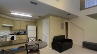 This two-floor space presents a practical and functional layout designed for comfortable everyday use. The image reveals a combined living and kitchen area on what appears to be a lower level, characterized by neutral beige walls and light-colored carpeting that contribute to a cohesive, understated aesthetic throughout.

The kitchen area is equipped with light wood cabinetry, a full-size white refrigerator, and a built-in microwave, offering ample storage and essential appliances. A compact dining area features a round granite-top table paired with traditional X-back chairs, providing an intimate space for meals.

The living area includes a black leather armchair, offering a casual seating option. Recessed vents and flush-mounted fluorescent lighting are integrated into the ceiling, ensuring consistent airflow and illumination throughout the space. A paneled utility access point is visible on one wall, indicating practical infrastructure within the unit.

A white interior door leads to an additional room, while a staircase in the background connects to the second floor, suggesting further living space above. Overall, the space maintains a clean, no-frills atmosphere focused on functionality and efficient use of space.