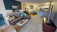 This thoughtfully designed 2-bedroom, 1-bathroom premium upgraded residence combines comfort and modern style in a seamless open-concept layout. The living area features plush carpeting and a welcoming atmosphere accented by bold teal and slate-gray wall tones, creating a striking visual contrast against neutral trim. A generously sized sectional sofa anchors the living space, complemented by a sleek coffee table and carefully curated décor that adds personality and warmth. The living and dining areas flow naturally into one another, with the transition marked by a shift to hardwood-style flooring in the dining zone. The dining area is appointed with a stylish table and upholstered chairs, creating an inviting setting for everyday meals or entertaining. A ceiling fan overhead adds both function and charm. Adjacent to the dining area, the kitchen features white cabinetry and modern appliances, offering a clean, bright contrast to the richer tones of the living spaces. The premium upgrade package is evident throughout, with cohesive finishes and elevated design choices that set this residence apart. The overall ambiance is contemporary and well-balanced, making excellent use of the available space to create a comfortable and visually engaging home.