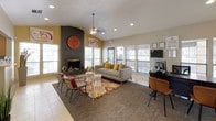This single-story community clubhouse offers a thoughtfully designed and energetic gathering space with a distinct personality. The main lounge area is anchored by a dramatic floor-to-ceiling dark brick fireplace, which serves as the room's focal point and rises to meet the vaulted ceiling. A bold, oversized red clock mounted above the firebox adds a striking decorative element. Vaulted ceilings throughout the space create an open, airy atmosphere, further enhanced by recessed lighting and a ceiling fan with a pendant-style fixture.

Natural light flows generously through multiple white-framed windows fitted with blinds, keeping the space bright and welcoming. The lounge is furnished with a contemporary gray sofa accented by mustard yellow pillows, wire-frame accent chairs, and a vibrant patterned area rug layered over tile and carpet flooring, lending the space a lively, eclectic character.

The entry area greets visitors with double glass-panel doors, a pendant light fixture, a starburst mirror, and a rustic wood credenza with a coffee station. Adjacent multi-purpose areas feature vaulted ceilings, colorful wall art, a refrigerator, and flexible seating, providing functional spaces suitable for work or casual gatherings.