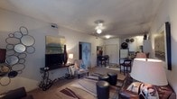 This single-floor residence is designed around an open-concept layout that seamlessly connects the living, dining, and kitchen areas, creating a cohesive and functional living environment. The living room serves as the central gathering space, featuring textured ceilings, a ceiling-mounted light fixture, and recessed HVAC vents. The room is anchored by an ornate wrought-iron media console topped with a flat-screen television, complemented by decorative accents including a tall vase and a table lamp. A patterned area rug with sweeping lines defines the seating zone, which includes a leather sofa, leather ottomans, and a cushioned accent chair. Decorative wall art adds character to the neutral-toned walls. The open layout flows naturally into a dining area outfitted with a dark wood high-top table and matching bar-height chairs, positioned adjacent to the kitchen. The kitchen is equipped with standard appliances, including a refrigerator, and features counter seating, offering both practicality and a social connection to the rest of the living space. Warm, neutral carpet throughout the main living areas contributes to the overall inviting ambiance of this well-organized and comfortably appointed home.
