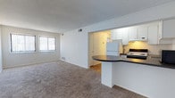 This single-floor, two-bedroom, one-bathroom unit offers a well-organized layout that maximizes its footprint. The open-concept design connects the living area to the kitchen, creating a cohesive and sociable space. The living room features wall-to-wall carpeting and a large window with vertical blinds, allowing natural light to brighten the room while providing privacy.

The kitchen is positioned centrally and opens to the living area via a peninsula counter with a dark laminate surface, offering additional workspace and a casual dining option. White cabinetry lines the kitchen walls, providing ample storage, while standard appliances including a refrigerator and electric range complete the setup. The kitchen floor transitions to hardwood-style flooring, distinguishing it from the carpeted living spaces.

The two bedrooms are situated on either side of the unit, each receiving natural light through individual windows. The shared bathroom is centrally located, offering convenient access from both bedrooms. The textured ceiling is consistent throughout the unit, adding a subtle design element to the otherwise clean, neutral interior. Overall, the space presents a practical and straightforward layout suited for efficient daily living.