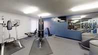 This fitness center offers a comprehensive workout environment designed to accommodate a wide range of exercise needs. The space is organized across a single floor and is divided into distinct functional zones, making it easy to navigate between cardio and strength training areas.

The cardio section is well-stocked with multiple treadmills, elliptical machines, and a stationary bike, all positioned on protective rubber mats atop carpeted flooring. A wall-mounted flat-screen television is conveniently placed for viewing during workouts. The strength training area features a multi-station cable machine, adjustable benches, and free weights, providing options for both resistance and functional training.

Large wall-to-wall mirrors line multiple sections of the room, visually expanding the space and allowing users to monitor their form during exercise. The walls are accented in a deep navy blue, contrasting cleanly with the white ceiling and trim to create a focused, energizing atmosphere. Flush-mounted fluorescent light panels illuminate the entire space evenly, ensuring a bright and welcoming environment at any time of day.

A window with vertical blinds allows for natural light in the strength area, while blue-painted interior doors provide access to additional sections of the facility. The textured ceiling and carpet throughout give the space a practical, low-maintenance finish suited to high-traffic use.