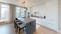 This elegantly designed high-rise residence offers a seamless blend of modern sophistication and comfortable living across a single thoughtfully laid-out floor. Floor-to-ceiling windows throughout flood the space with natural light and frame sweeping panoramic views of the city skyline, lakefront, and waterway beyond.

The gourmet kitchen is a standout feature, outfitted with crisp white shaker cabinetry extending to the ceiling, dark stone countertops, a professional gas cooktop, double wall ovens, and a large central island with bar seating — all illuminated by a striking contemporary pendant light. Adjacent to the kitchen, the dining area enjoys generous window exposure with waterfront views and light hardwood flooring that flows throughout the main living spaces.

The living room exudes a calm, refined ambiance with textured grasscloth wall coverings, large-format windows overlooking the lake, and an open, welcoming layout. A versatile secondary room, painted in a soft steel blue, features a built-in shelving and workspace system concealed behind sliding doors — ideal as a home office or guest space.

The primary bedroom offers serene waterfront views through expansive windows and is complemented by a luxurious en-suite bathroom featuring marble herringbone tile flooring, a double vanity with backlit mirrors, and a glass-enclosed walk-in shower. A secondary bathroom echoes the same refined aesthetic with herringbone tile and a classic white vanity.