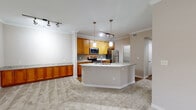 Located at 9600 Coit Rd in Plano, Texas, this single-floor residence offers a well-designed, open-concept layout finished with neutral tones throughout. Crown molding accents the main living areas, which feature plush carpet flooring and generous natural light from multiple windows fitted with blinds.

The kitchen serves as a central hub of the home, boasting warm honey-toned cabinetry, light granite countertops with a subtle tile backsplash, and a full suite of stainless steel appliances including a range, over-the-range microwave, and refrigerator. A large center island with an integrated sink and pendant lighting above provides additional prep and seating space. Track lighting adds supplemental illumination throughout the kitchen and living area.

The spacious living room includes built-in shelving along one wall, a ceiling fan, and a glass door providing direct outdoor access. An adjacent room also offers exterior access through a windowed door, allowing ample natural light and outdoor views.

The bathrooms feature full tub-shower combinations with white tile surrounds and wood-look flooring. One bathroom connects directly to a walk-in closet equipped with wire shelving, offering practical storage. Additional built-in shelving and closet storage are found throughout the unit, enhancing overall functionality.