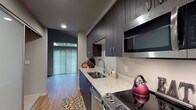 This single-floor unit showcases a thoughtfully designed layout with modern finishes throughout. The galley-style kitchen is a standout feature, equipped with dark espresso cabinetry extending to the ceiling, light granite countertops, a stainless steel sink, and a full suite of stainless steel appliances including a smooth-top range, over-the-range microwave, dishwasher, and refrigerator. Recessed lighting illuminates the kitchen, which connects seamlessly to the rest of the unit.

The living area benefits from vaulted ceilings that enhance the sense of openness, while large sliding glass doors and an additional window flood the space with natural light and provide views of surrounding greenery. Light wood-look flooring runs consistently throughout the unit, creating visual continuity between rooms.

The bathroom is clean and functional, featuring a dark wood vanity with a granite-topped sink, a large frameless mirror, and well-placed lighting. The space maintains the same light flooring found throughout the unit.

A short hallway connects the main living areas to the entry, where a utility panel is conveniently located. The overall design balances modern aesthetics with practical functionality, offering a cohesive and comfortable living environment.