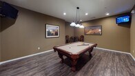 This single-floor amenity space offers a thoughtfully designed layout with a diverse range of recreational and functional areas. A wide, tile-floored corridor connects the various rooms, creating an open and navigable flow throughout the facility. The hallway is lined with glass-paneled doors and recessed lighting, lending a clean, welcoming atmosphere.

The standout game room features rich hardwood-style flooring and warm earth-toned walls adorned with framed landscape artwork. A full-size billiard table with ornate carved legs takes center stage beneath an elegant multi-pendant chandelier, while a seating area in the corner adds a social touch. The fitness center, accessible directly from the main corridor, is equipped with cardio machines and workout equipment beneath a ceiling fan, with natural light filtering in through a large window.

A cozy media lounge offers a spacious sectional sofa, soft sconce lighting, and wood-look flooring — ideal for movie screenings or casual gatherings. An adjacent room, visible through a glass-inset door, features a warm fireplace and comfortable seating, providing a relaxed retreat. Throughout the space, recessed lighting, neutral color palettes, and tile and wood-look flooring combine to create a cohesive, comfortable, and inviting environment suited for a variety of activities.