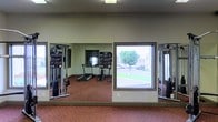 Located at 20 N Hill Pky in Jackson, Mississippi, this single-story fitness center offers a well-equipped workout space designed to support a variety of exercise routines. The room features large floor-to-ceiling mirrors lining the walls, creating a sense of openness and allowing users to monitor their form during workouts. The space is furnished with a treadmill and a stair climber for cardiovascular training, as well as multi-function cable and weight machines to accommodate strength training exercises. The flooring consists of durable dark red rubberized material, well-suited for high-traffic fitness use. Natural light fills the room through a prominent window that also provides views of the surrounding outdoor greenery and the adjacent building exterior. The neutral-toned walls and overhead lighting contribute to a clean, functional atmosphere. The layout is open and uncluttered, allowing for comfortable movement between equipment. Overall, this fitness center provides a practical and accessible environment for residents or users seeking a convenient on-site exercise facility.