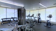 This fitness center offers a comprehensive workout environment designed to accommodate a variety of exercise needs. The space is bright and open, benefiting from large windows fitted with roller shades that allow natural light to filter in while maintaining a comfortable atmosphere for exercise.

The cardio section is well-appointed, featuring multiple treadmills and an elliptical trainer, each equipped with individual display consoles for tracking workout metrics. Complementing the cardio equipment is a multi-station cable and weight machine, which provides users with options for a wide range of strength-training exercises. A dedicated free weights area with a dumbbell rack rounds out the strength training offerings.

The flooring throughout the facility is a durable, dark rubber material — ideal for both heavy equipment and high-impact activity. Overhead fluorescent lighting ensures the space remains well-lit throughout all hours. The clean, modern aesthetic of the room is consistent with a professional fitness environment, keeping the focus on functionality and utility. Ventilation is supported by ceiling vents, contributing to a comfortable training atmosphere. Overall, this fitness center is thoughtfully organized to provide users with an efficient and fully functional workout experience.