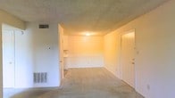 This single-floor unit offers an open and straightforward layout centered around a carpeted living area. The space is finished with neutral white walls and a textured ceiling that adds subtle dimension overhead. A flush-mount ceiling light provides central illumination, complemented by HVAC vents positioned along the walls for climate control.

Visible from the main living area, a partially open kitchen sits to one side, featuring white cabinetry and countertop space. The transition between the living and kitchen areas is defined by a subtle change in flooring, with the kitchen appearing to have a hard surface contrasting the soft carpet underfoot in the main room.

Two doors are visible from the living space — one appearing to lead to an exterior entry and another to an adjacent room or closet — contributing to the unit's functional flow. The overall layout feels compact yet efficient, with each element serving a clear purpose. The warm, neutral palette throughout gives the space a clean and move-in-ready appearance, suitable for straightforward, comfortable everyday living.