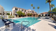 Located at 8092 W Paradise Ln in Peoria, Arizona, this outdoor amenity space embodies the spirit of resort-style living in the Sonoran Desert. The centerpiece of the area is a generously sized swimming pool with crystal-clear blue water, framed by a wide concrete pool deck furnished with rows of lounge chairs and patio umbrellas — ideal for relaxing under the Arizona sun. Towering palm trees punctuate the landscape, providing natural shade and a tropical aesthetic against the backdrop of terracotta-roofed residential buildings. A covered cabana anchors one end of the pool deck, constructed with stone-accented pillars and a tiled roof, offering a shaded gathering space equipped with seating and an outdoor bar or service counter. Adjacent to the pool area, a putting green with hammock-style loungers provides an additional recreation zone, blending leisure and activity in a compact, well-designed layout. The surrounding buildings feature a Southwestern architectural style with stucco exteriors, pitched tile roofs, and stone detailing that harmonize with the natural desert environment. Mature trees and desert-adapted plantings soften the hardscape, contributing to an inviting, verdant atmosphere. The overall setting is bright, open, and well-appointed, reflecting a community designed with outdoor living and recreation at its core.