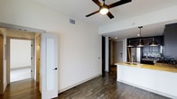 This single-floor space features an open-concept layout that seamlessly connects the living area to the kitchen, creating a functional and cohesive flow throughout. The flooring is dark hardwood-style planks, which run continuously across the space, lending warmth and a contemporary feel to the interior.

The living area is bright and clean, with white walls and elevated ceilings fitted with a multi-blade ceiling fan with integrated lighting, enhancing both air circulation and ambient light. A paneled interior door with a dark hardware finish adds a classic detail to the space.

The kitchen is positioned toward the rear and features dark cabinetry paired with a light stone countertop island that includes a built-in sink. Pendant lighting in dark metal finishes hangs above the kitchen area, complementing the cabinetry. A stainless steel refrigerator is visible, indicating modern appliance finishes throughout.

The entryway, visible beyond the kitchen, is finished with a solid door and recessed lighting, completing the overall modern and well-appointed character of this single-floor space.