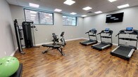 This fitness center offers a clean, modern environment designed to support a well-rounded workout routine. The space is finished with rich hardwood-style flooring throughout, complemented by neutral gray walls and a drop ceiling fitted with large panel LED lights that provide consistent, even illumination across the entire floor.

The cardio area features three treadmills with individual digital consoles, positioned along one wall beneath a wall-mounted flat-screen television. On the opposite side of the room, a recumbent stationary bike and an adjustable workout bench provide options for low-impact cycling and strength training. A multi-functional cable machine rounds out the strength equipment, offering a versatile range of resistance exercises.

Horizontal windows along one wall admit natural light and offer views of the surrounding brick exterior, adding an open feel to the space. A water cooler station and a small storage cabinet with sanitation supplies are conveniently located near the entrance, supporting hygiene and hydration during workouts. Additional amenities include a dedicated entry door with frosted glass panels. The overall layout provides ample open floor space for stretching or functional movement, making this fitness center a practical and comfortable facility for users of varying fitness levels.