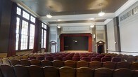 This single-floor auditorium exudes a timeless, classical character defined by its refined architectural detailing and functional performance layout. The centerpiece of the space is a proscenium-style stage framed by rich red curtains, providing a dramatic focal point for performances, presentations, or lectures. Flanking the stage are symmetrically placed doorways with decorative pediment surrounds, adding a formal elegance to the overall design.

Above the stage, an ornate decorative frieze runs the width of the hall, featuring classical relief-style figures that enhance the venue's historic aesthetic. The walls are finished with clean white plasterwork accented by additional decorative panels, contributing to the room's dignified atmosphere. Pendant light fixtures hang from the ceiling, complementing the ambient lighting provided by wall-mounted units throughout the space.

Rows of burgundy upholstered fixed seating fill the auditorium floor, offering generous capacity for audiences. Tall windows along the side walls allow natural light to filter into the hall. The overall ambiance is one of understated grandeur, making this venue well-suited for theatrical productions, community events, and formal gatherings.