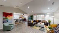 Located at 733 29th St NW in Roanoke, Virginia, this single-floor community space offers a thoughtfully designed interior that balances functionality with a warm, inviting atmosphere. The open-concept layout is anchored by soaring vaulted ceilings that amplify the sense of space throughout, while recessed lighting and large windows with blinds allow abundant natural light to fill the interior.

The main lounge area is generously proportioned, featuring a large sectional sofa accented with colorful throw pillows, an upholstered ottoman, and a vibrant area rug that adds energy to the room. Tall floor lamps and potted greenery contribute to the welcoming ambiance. Multiple exterior doors provide easy access and enhance the flow of natural light.

Adjacent to the lounge is a reception and service counter area equipped with ample counter space, a sink, cabinetry, and a wall-mounted display screen — ideal for administrative or community service functions. A breakfast bar with bar-height seating and a ceiling fan connects this zone to a casual dining area.

The entry foyer features vaulted ceilings, bold abstract artwork on the walls, a console cabinet, and a comfortable accent chair, creating a polished first impression. Throughout the space, wood-look flooring ties the areas together cohesively, while the colorful décor scheme adds a lively, community-centered character to the overall environment.