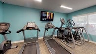Located at 2738 Roosevelt Blvd in Largo, Florida, this fitness center offers a well-organized, single-floor workout facility designed to support a full range of exercise routines. The space is characterized by vibrant teal-painted walls, neutral multi-tone carpet tiles, and bright overhead fluorescent lighting that creates an energetic and welcoming atmosphere throughout.

The main workout area is generously sized and divided into distinct functional zones. A dedicated cardio section houses multiple Matrix treadmills and elliptical cross-trainers, complemented by a wall-mounted flat-screen television for entertainment during workouts. Adjacent to this area, a strength training zone features a multi-station cable and weight machine, along with a bench press unit for targeted resistance training.

A separate section of the floor is devoted to functional fitness and flexibility, equipped with a rack of stability balls in assorted sizes, resistance bands, medicine balls, and exercise mats — ideal for stretching, balance, and core training. A dual-level water fountain with a bottle-filling station is conveniently positioned along one wall, alongside a paper towel dispenser and waste receptacle for hygiene and convenience.

The facility also includes a storage room with a glass-panel door, providing organized space for additional equipment. Windows along the perimeter allow natural light to filter in, enhancing the open feel of the space. Safety features, including a fire extinguisher and illuminated exit signage, are clearly visible throughout.