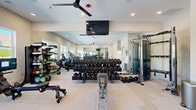 Located at 235 McDonough Rd in McDonough, GA, this fitness center is a comprehensively outfitted workout facility designed to support a full range of exercise routines. The space features light-toned flooring throughout, recessed lighting, and ceiling fans that contribute to a bright and comfortable environment. Large windows along one wall allow natural light to fill the room, offering views of the surrounding outdoor area and creating an open, airy atmosphere. Floor-to-ceiling mirrors line multiple walls, visually expanding the space and allowing users to monitor their form during workouts. The cardio zone includes treadmills, an elliptical trainer, and stationary cycling bikes, each equipped with individual display screens. The free weights section features a multi-tiered dumbbell rack with an extensive range of weights, accompanied by a flat bench for versatile training. A Matrix power rack with a barbell and adjustable bench supports strength and powerlifting exercises. Additional functional training equipment, including kettlebells, medicine balls, and cable machines, rounds out the offerings. A ceiling-mounted flat-screen television enhances the workout experience. Near the entrance, a built-in counter with a compact refrigerator and pendant lighting provides a convenient refreshment station, completing this well-appointed fitness facility.