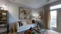 This thoughtfully designed single-floor space exudes a modern, sophisticated ambiance anchored by a welcoming living area. The room is finished in soft, neutral gray tones that create a calm and cohesive atmosphere throughout. A plush light-gray sofa serves as the centerpiece, adorned with an artful mix of patterned and solid throw pillows in warm earth tones. Above the sofa, a large-scale abstract artwork commands attention, adding a bold artistic focal point to the space. A gold-framed mirror on the adjacent wall reflects light and enhances the sense of depth. The living area is further complemented by a sleek glass-top coffee table styled with decorative candles and a wooden tray, adding layers of texture and warmth. A tall table lamp provides warm ambient lighting, while floor-length drapes in a soft taupe frame the window, lending an air of elegance. The flooring features a richly patterned area rug that grounds the seating arrangement and introduces a touch of color. Subtle architectural details, including clean lines and a well-proportioned ceiling with a discreet HVAC vent, speak to the thoughtful construction of the space. Overall, the interior strikes a refined balance between comfort and contemporary style.