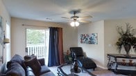 This single-floor space offers a warm and inviting atmosphere designed for comfortable everyday living. The living room serves as the heart of the home, showcasing a textured ceiling with a classic multi-blade ceiling fan that provides both lighting and air circulation. Soft, neutral-toned walls create a versatile backdrop, complemented by decorative wall art that adds a touch of color and personality to the room.

Natural light flows generously into the living area through a large sliding glass door dressed with floor-length curtains in rich navy and warm brown tones, offering a pleasing view of a private outdoor balcony and lush greenery beyond. The balcony, enclosed by a white railing, extends the living space outdoors and provides a pleasant spot for relaxation.

The living room is furnished with plush, dark upholstered seating, including a tufted leather chaise lounge that adds an elegant focal point, alongside a glass-top coffee table with ornate iron legs. Carpeted flooring throughout contributes to the cozy ambiance. Subtle decorative touches, including potted plants and accent pieces, round out the space, creating an environment that is both functional and aesthetically inviting.