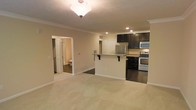 This single-story space showcases an open-concept design that seamlessly connects the living area to the kitchen. The living area features plush, light-toned carpeting and neutral beige walls, creating a warm and inviting atmosphere. Crown molding runs along the ceiling perimeter, adding a refined architectural detail, while a decorative pendant light fixture serves as a focal point overhead.

The kitchen is distinguished by dark espresso cabinetry that contrasts sharply with the lighter walls and countertops. A peninsula with bar-height seating provides a casual dining option and acts as a natural divider between the kitchen and living areas. Stainless steel appliances, including a refrigerator, microwave, and range, are integrated throughout. The kitchen features dark hardwood-style flooring, offering a clean transition from the carpeted living space.

Recessed lighting in the kitchen ensures functional illumination, complementing the overall lighting scheme. A hallway visible from the living area leads to additional rooms, including what appears to be a bathroom. The consistent use of crown molding and neutral color palette ties the entire space together cohesively.
