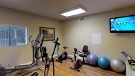 This single-floor fitness center offers a practical and well-organized space for exercise and physical wellness. The room features warm-toned walls and durable hardwood-style flooring, creating a clean and inviting environment for workouts. Overhead, a large flush-mount fluorescent light fixture provides bright, even illumination throughout the space. The fitness area is equipped with a variety of cardio machines, including an elliptical trainer, an upright stationary bike, and a recumbent bike, catering to users of different fitness levels and preferences. Exercise balls in multiple sizes are stored along one wall, adding versatility for stretching and balance training. A framed landscape painting adorns the wall, adding a subtle decorative touch to the otherwise functional space. A posted notice on the adjacent wall outlines fitness center guidelines, indicating that this facility serves a shared or community setting. The room is well-ventilated with a ceiling vent, and the layout allows for comfortable movement between equipment. Overall, the space is efficiently designed to deliver a focused and accessible fitness experience.